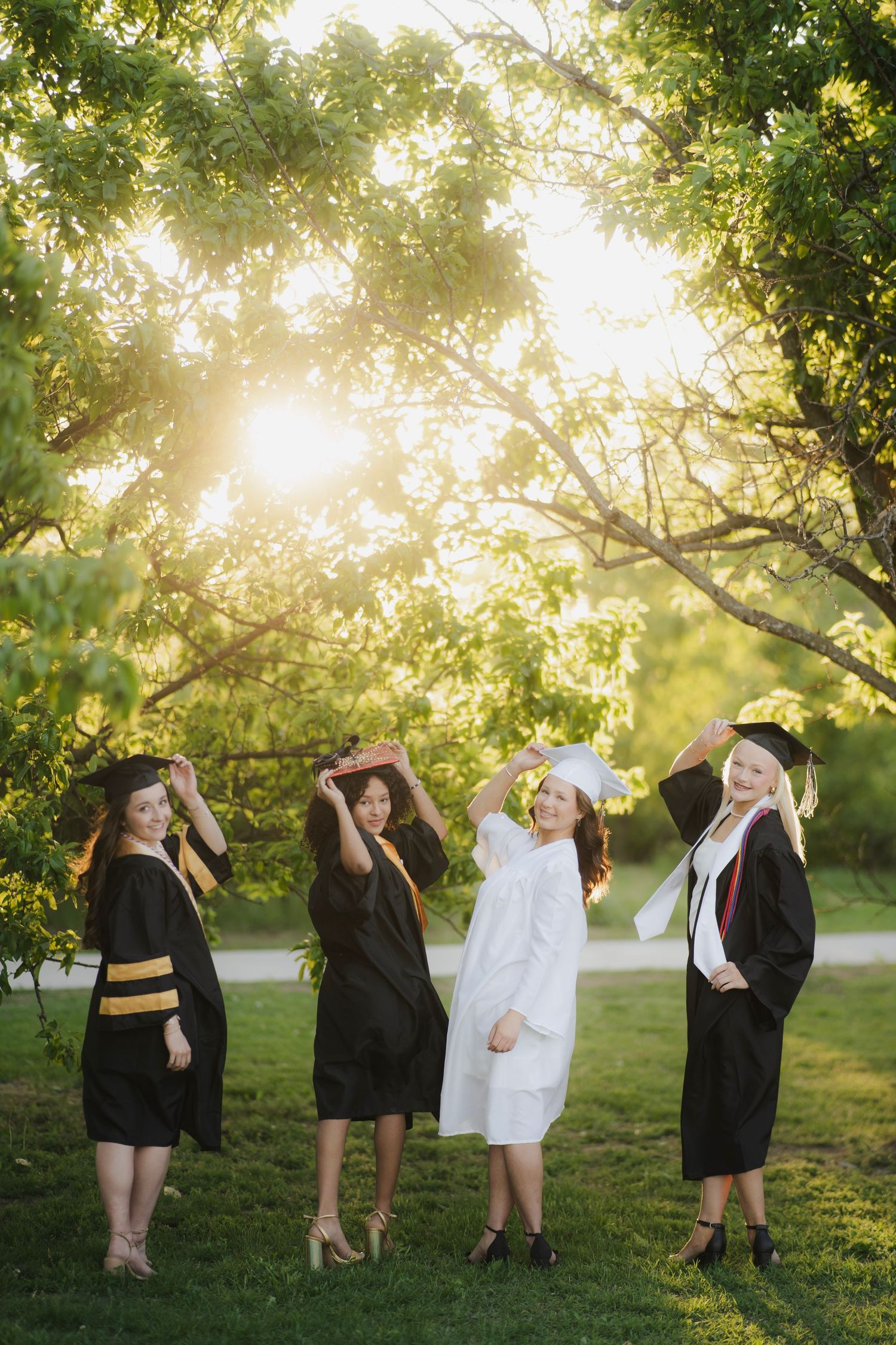 Four female graduates in caps and gowns playfully toss their hats in a sunlit park setting.