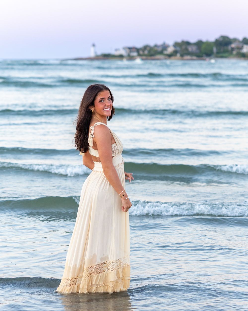 High school senior cream lace dress stands in the ocean at sunset at Wingaersheek Beach in Gloucester, MA for yearbook portrait session.