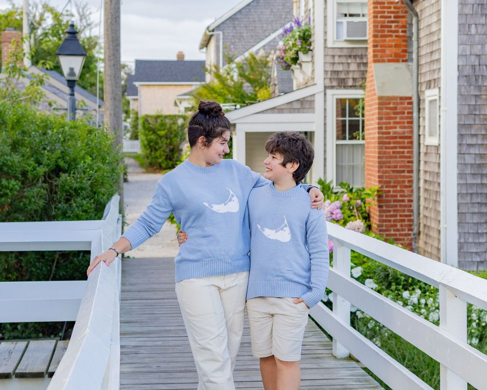 Siblings in matching blue sweaters hugging and smiling on the pedestrian bridge in Sconset on the island of Nantucket for family photos.