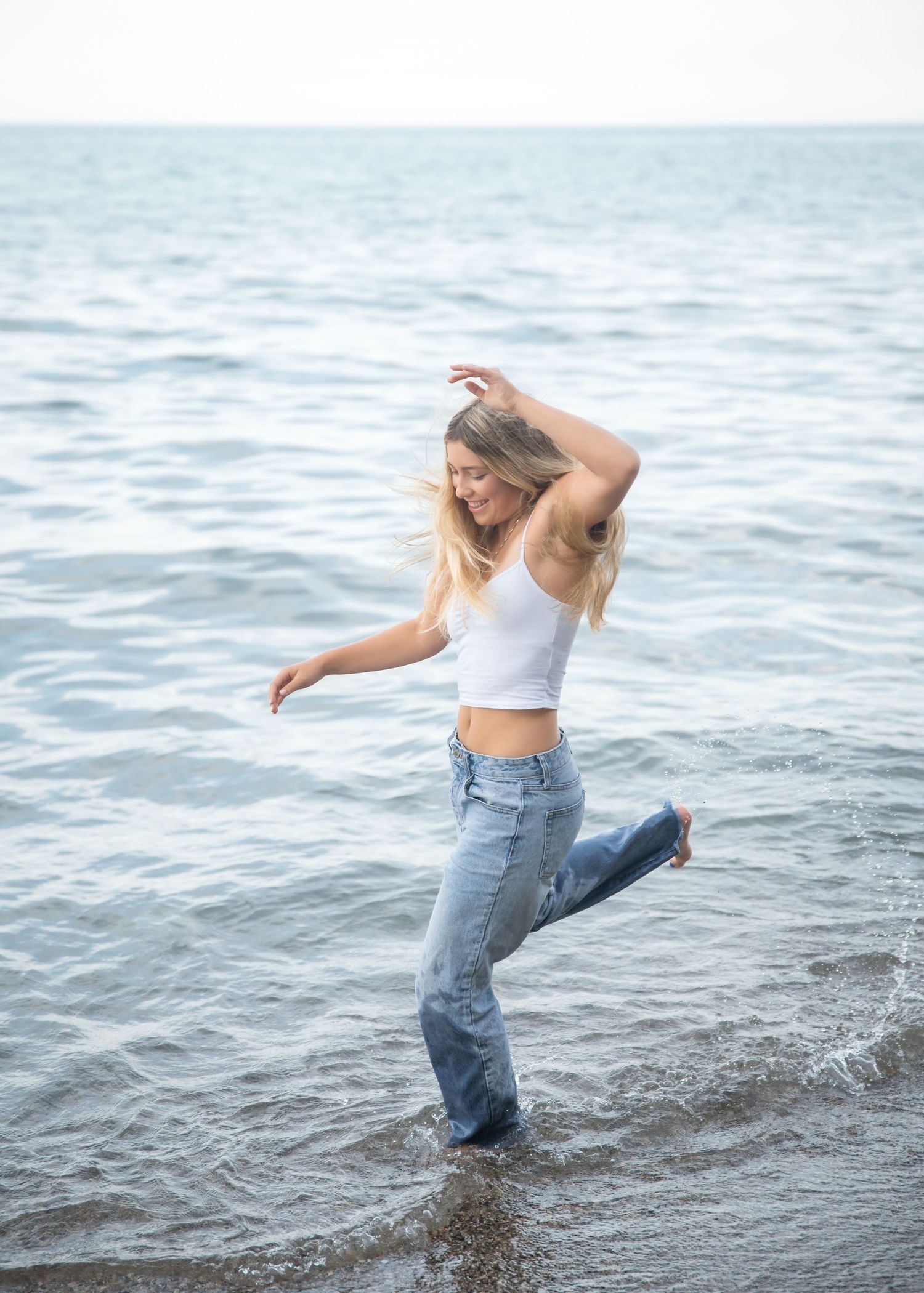 Blonde woman in white crop top and jeans dancing in ocean waves on a beach.
