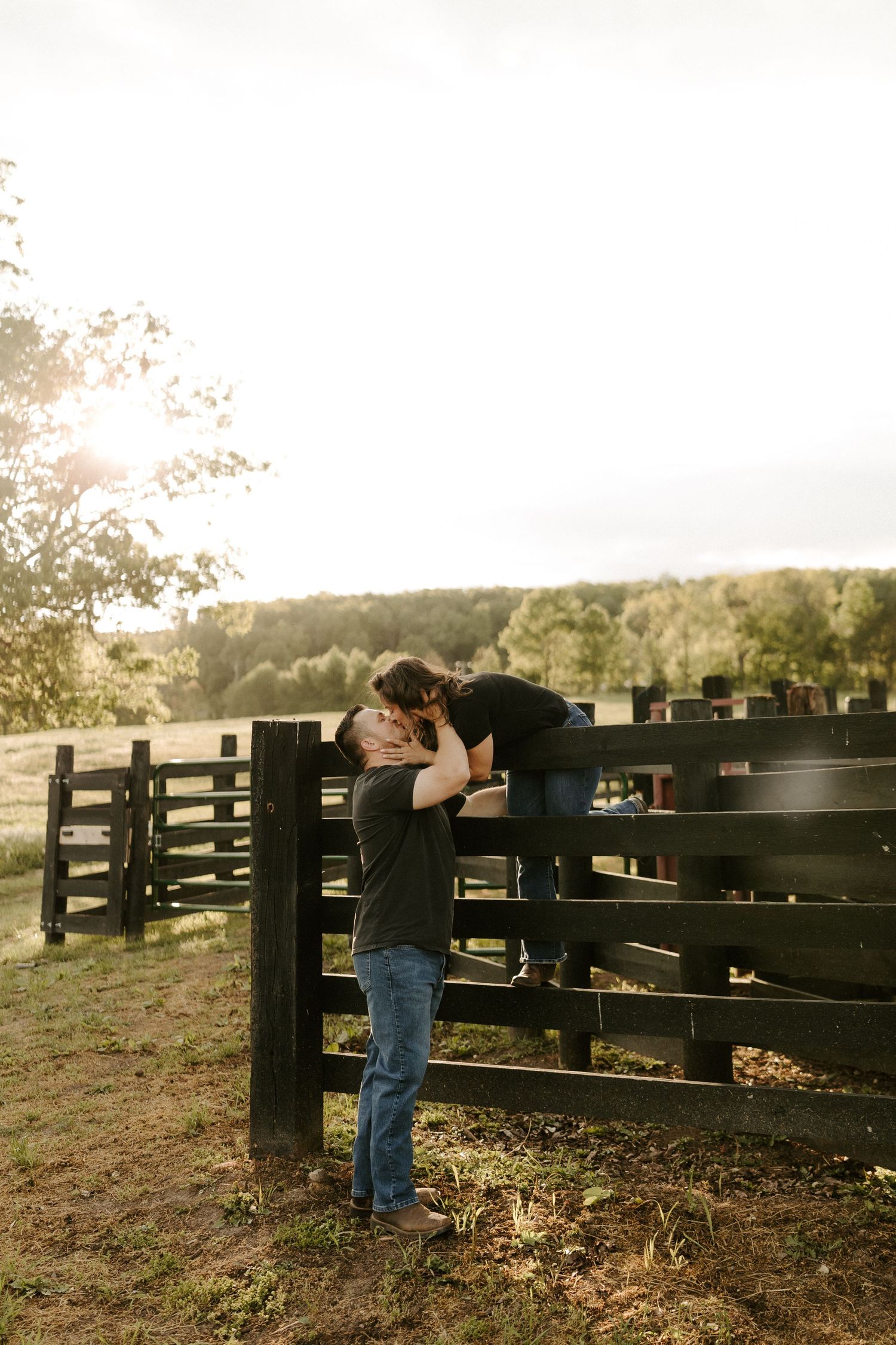 Man lifts woman over rustic wooden fence in sunny countryside setting.