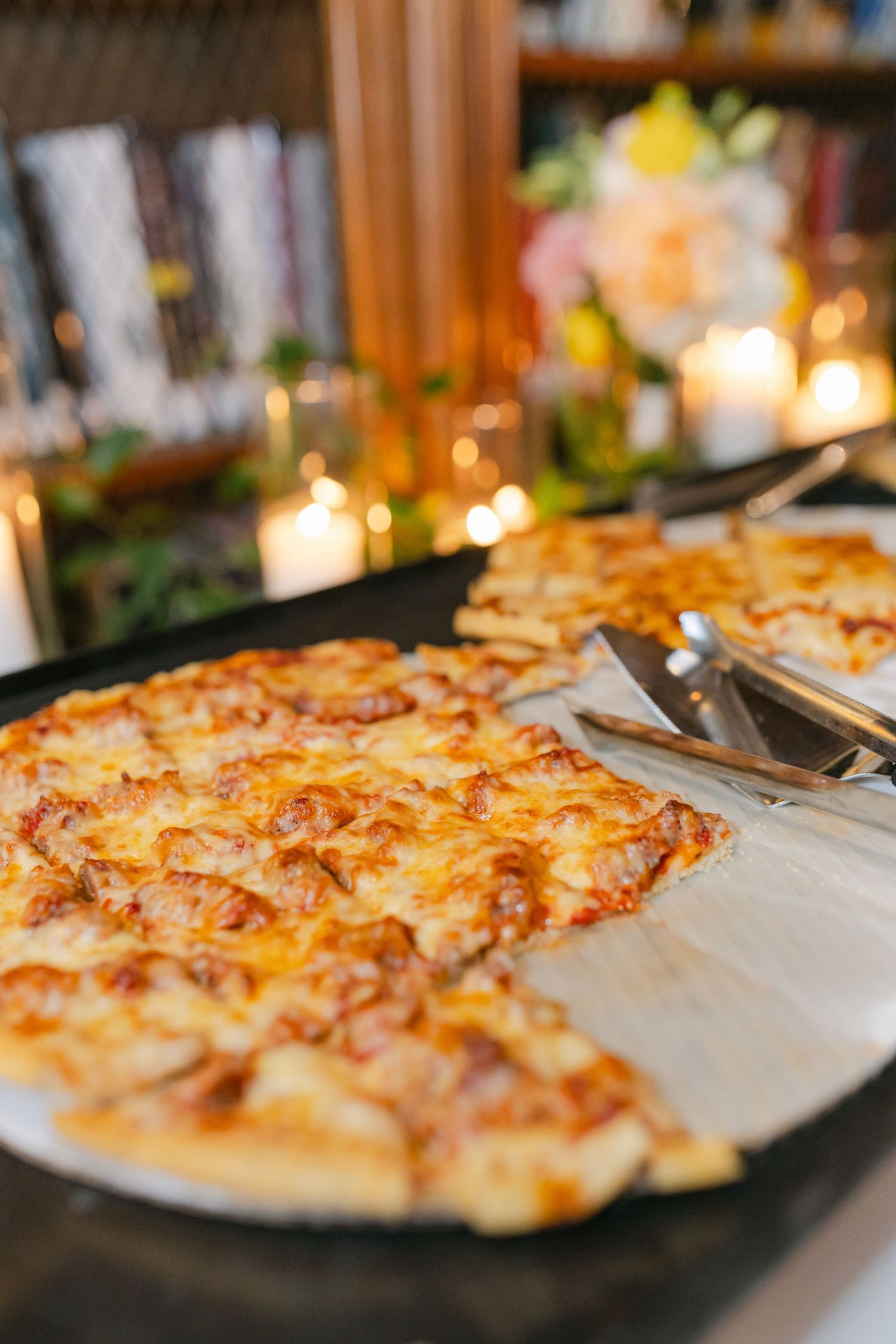 Cheesy pizza on a black pan with a server utensil, set on a buffet table with candles and floral decor.