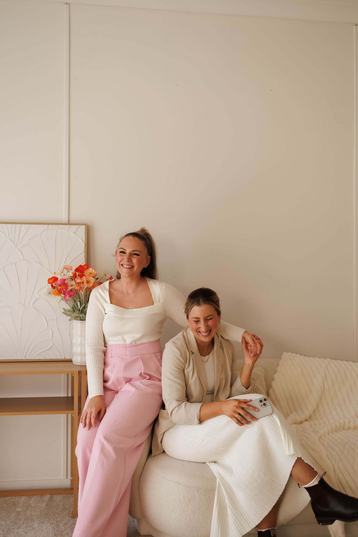 Two smiling women sit together in a bright, minimalist room with pink florals and neutral decor.