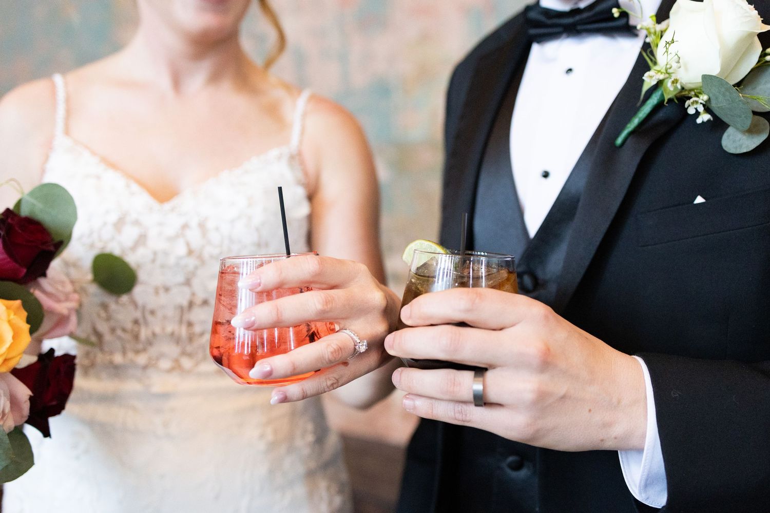 Bride and groom holding cocktails at wedding reception, wearing white lace gown and black tuxedo.