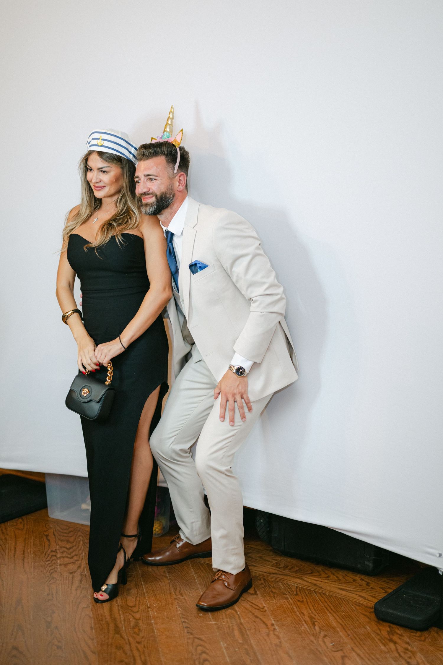 Couple in formal attire posing playfully, woman in black gown with sailor hat, man in beige suit with party hat.