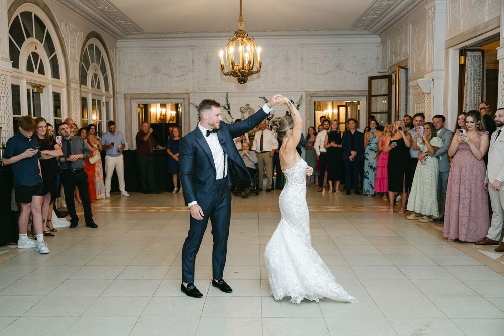 Bride and groom share first dance in elegant ballroom with chandelier and guests applauding in background.