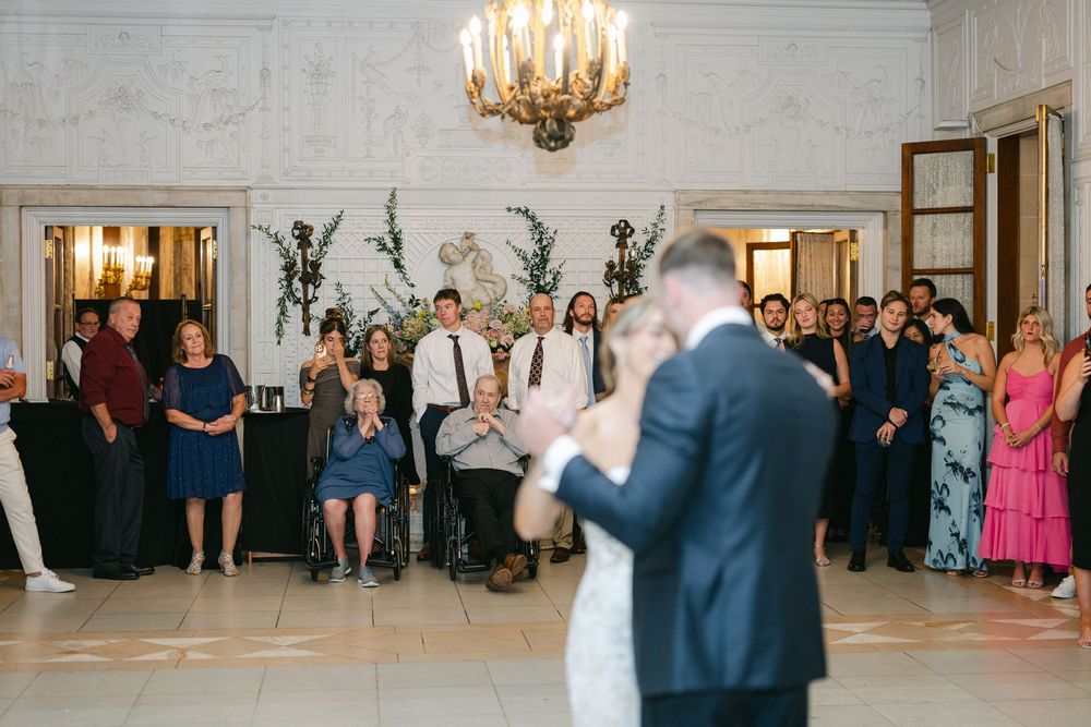 Bride and groom share first dance in elegant ballroom while wedding guests watch and celebrate around them.