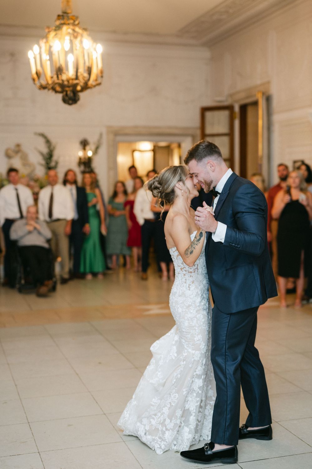 Bride and groom share first dance in elegant ballroom with chandelier and guests watching at wedding reception.