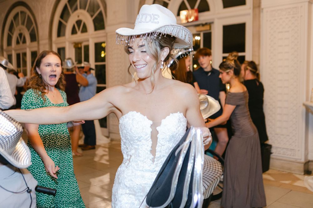 Bride in strapless lace gown and white cowboy hat dancing joyfully at wedding reception with guests celebrating.