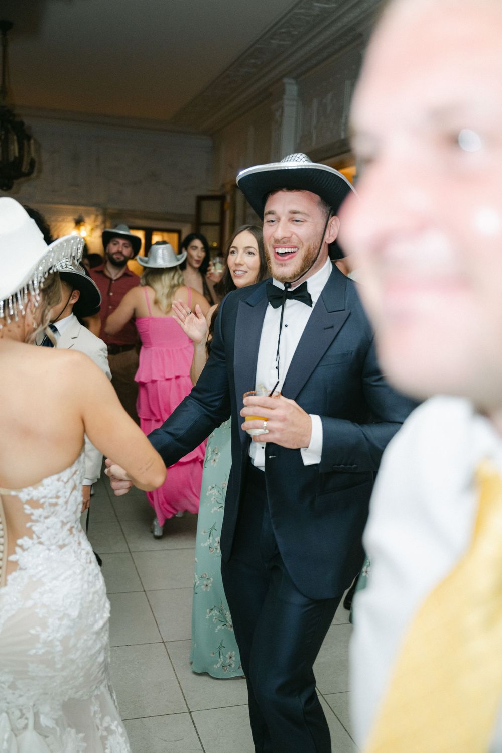 A groom in a black tuxedo and cowboy hat laughs joyfully while dancing with guests at a lively wedding reception.