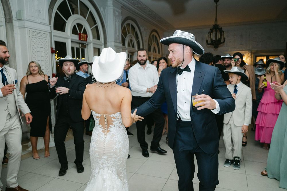 Bride and groom dancing in cowboy hats at wedding reception while guests watch in elegant venue.