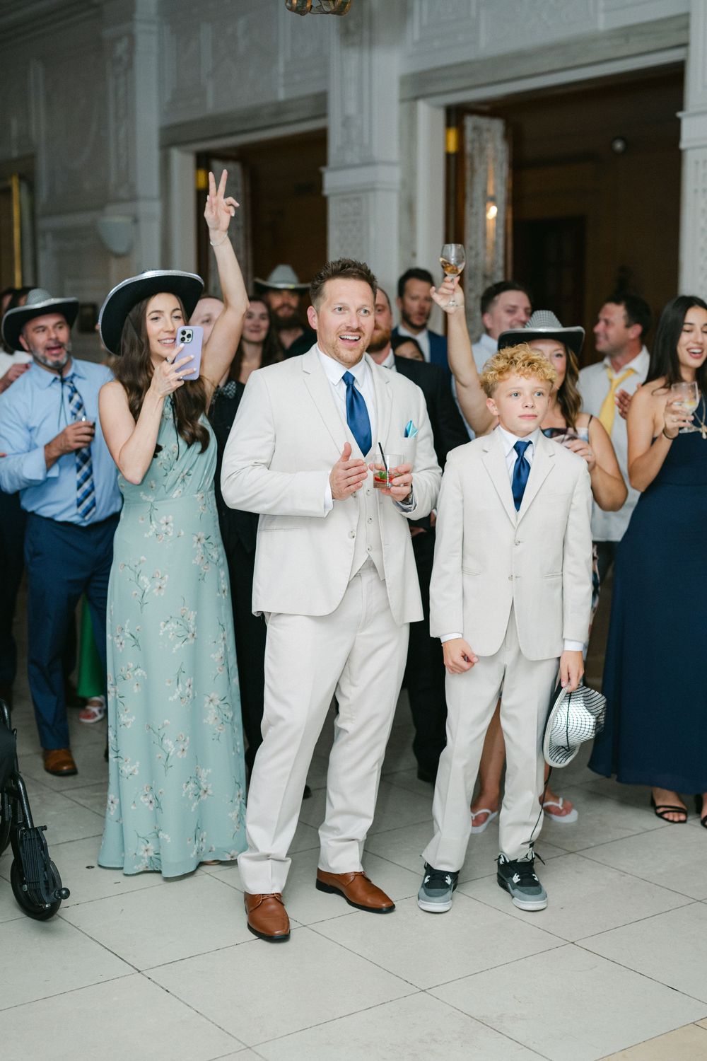 A man and boy in matching white suits with blue ties smile at a wedding reception surrounded by celebrating guests.