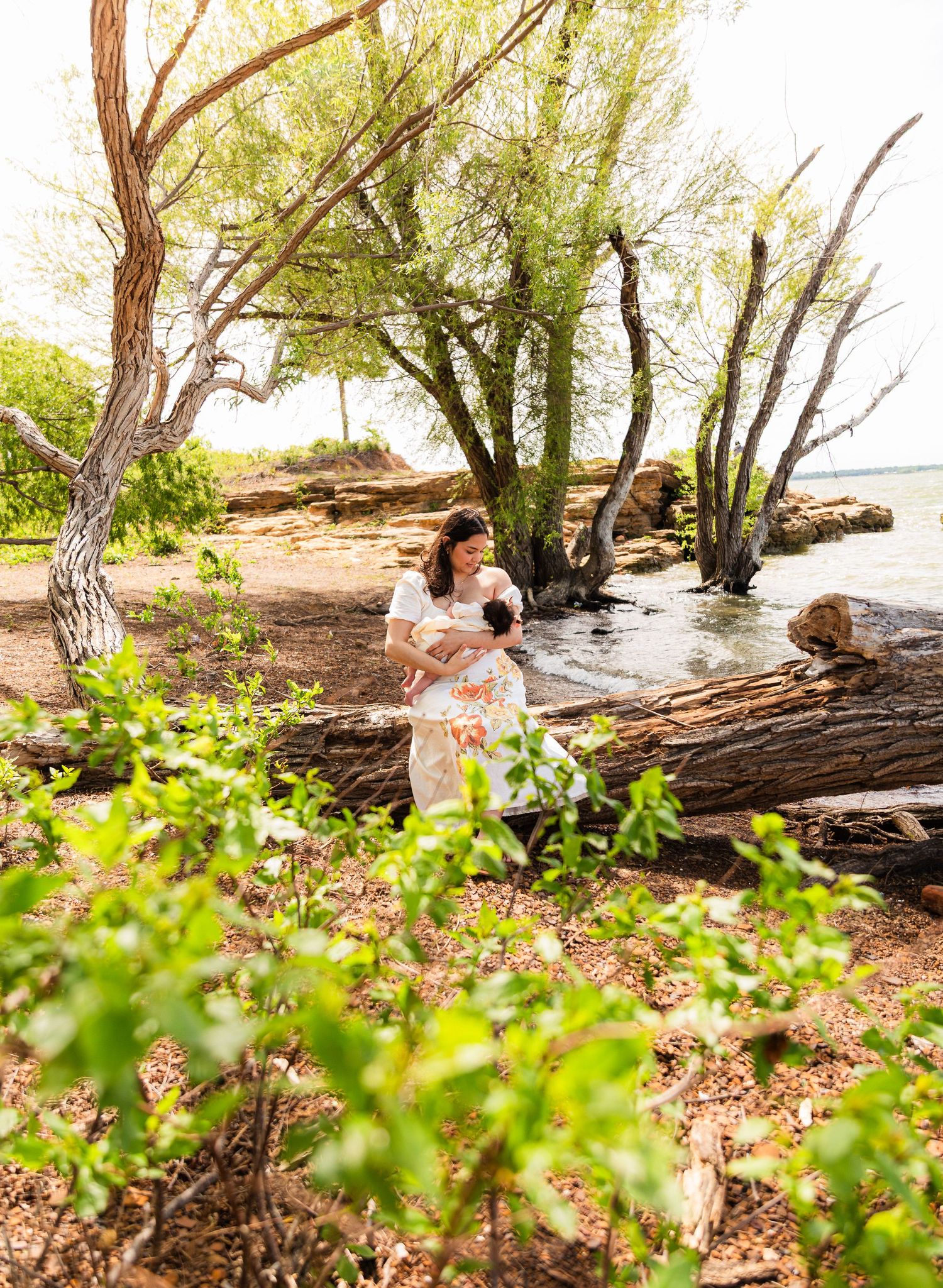 A woman in a floral dress holds a baby near a rocky shoreline surrounded by lush green trees.