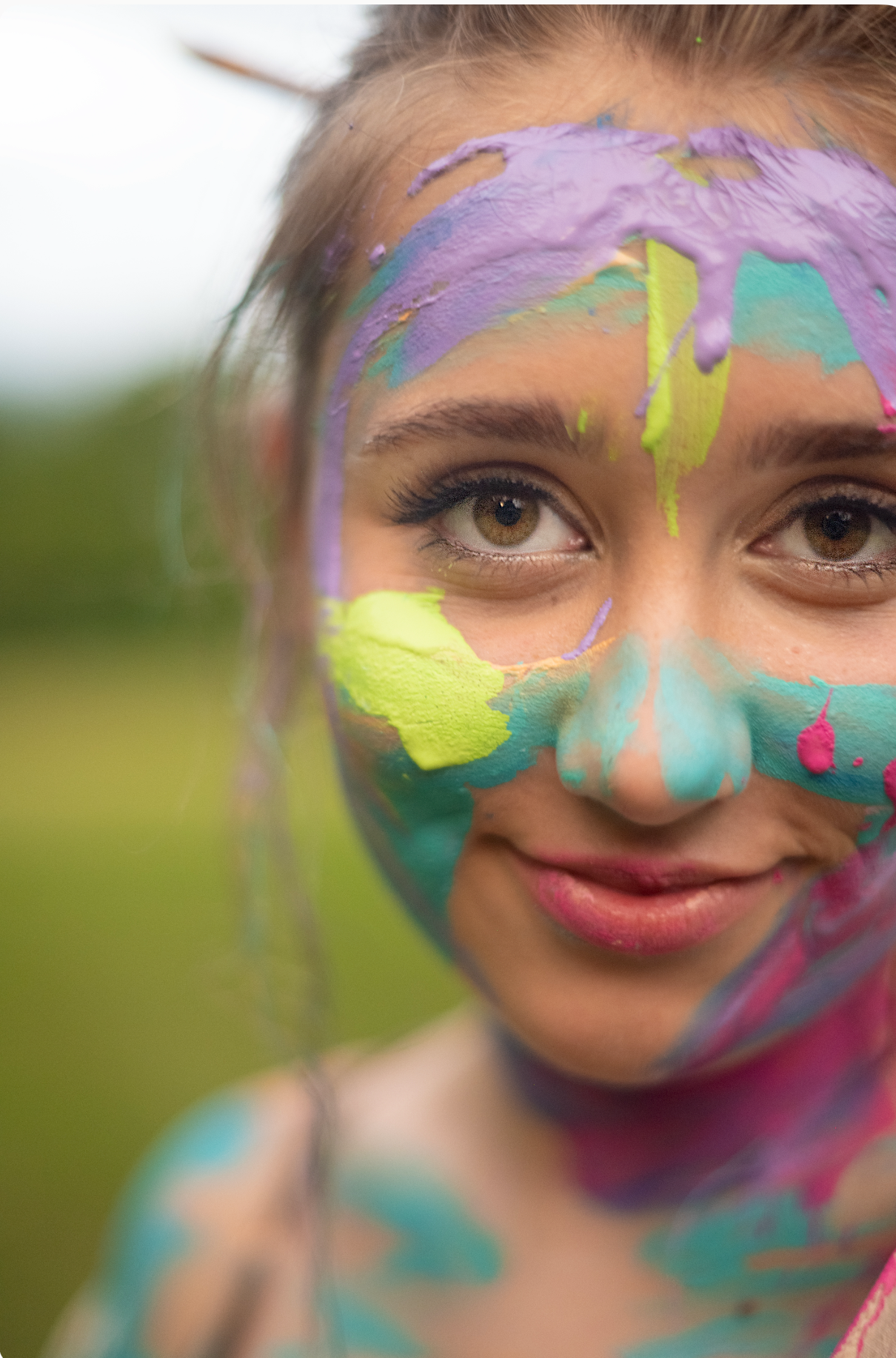 Young girl with colorful paint covering her face smiling outdoors, featuring purple, teal, yellow, and pink hues.