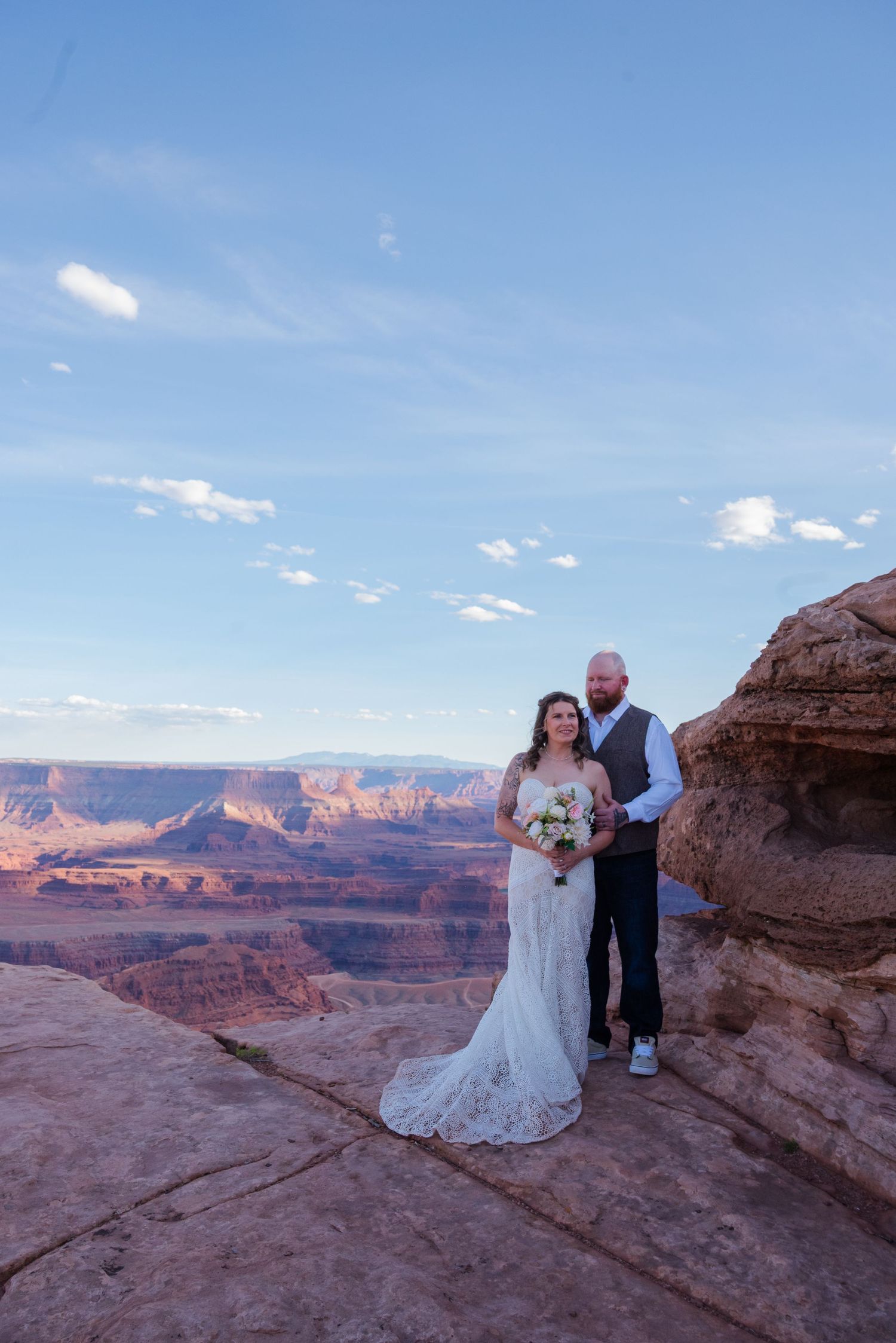 Bride and groom pose on a dramatic canyon overlook with sweeping red rock landscape under a blue sky.