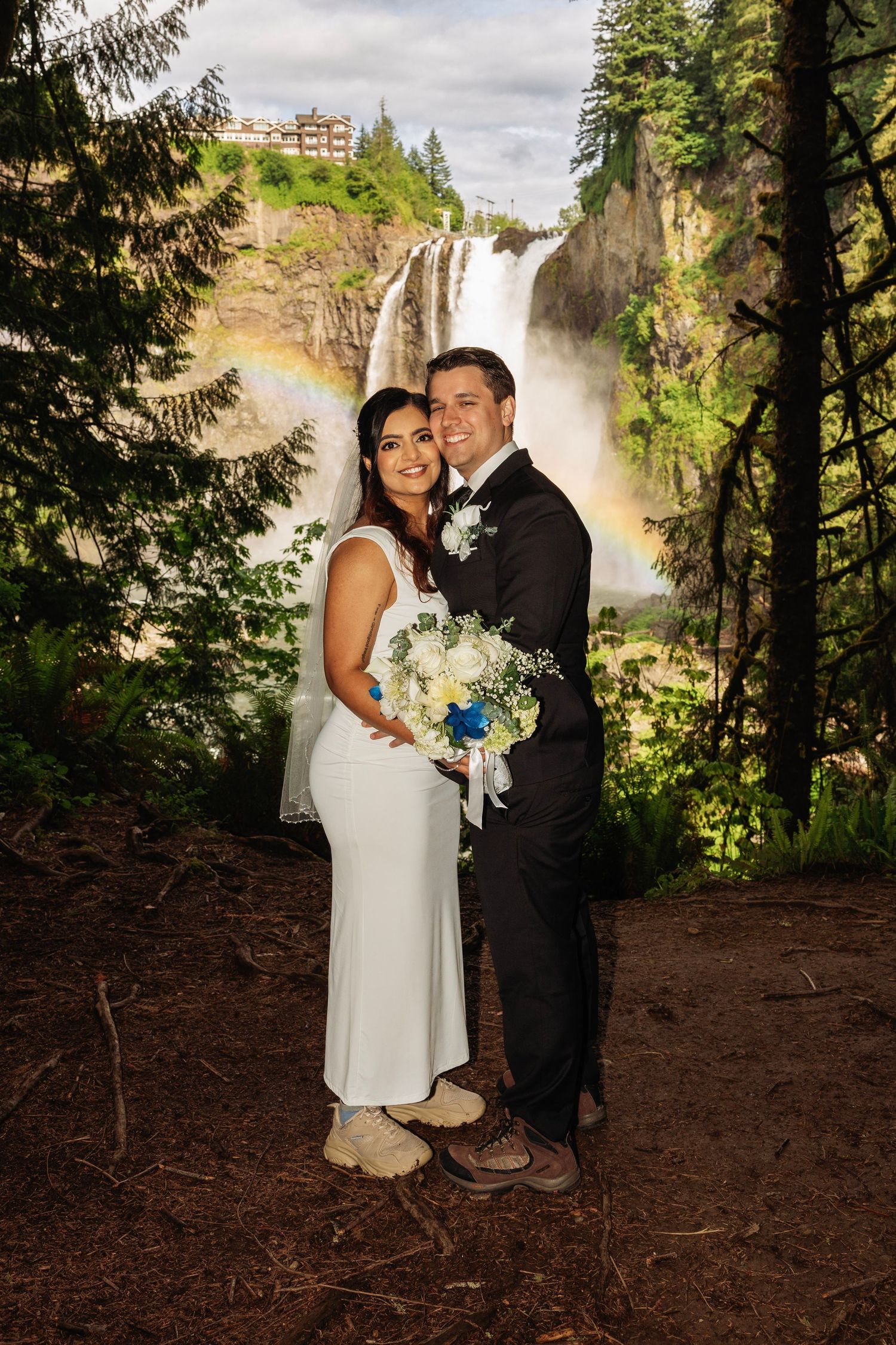 Bride and groom pose before Snoqualmie Falls with a rainbow, bride holding white bouquet in lush forest setting.