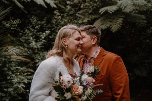 Two brides share a romantic forehead touch outdoors, one in a white fuzzy sweater holding a boho bouquet, the other in a rust suit.