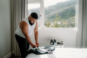Man in white tank top folding suit jacket on bed near window with mountain view and dress shoes nearby.