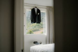 A wedding suit jacket with boutonniere hangs on a window in a bright bedroom with scenic green views outside.