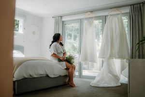 Bride in white robe sits on bed holding bouquet, wedding gown hanging nearby in bright bedroom.