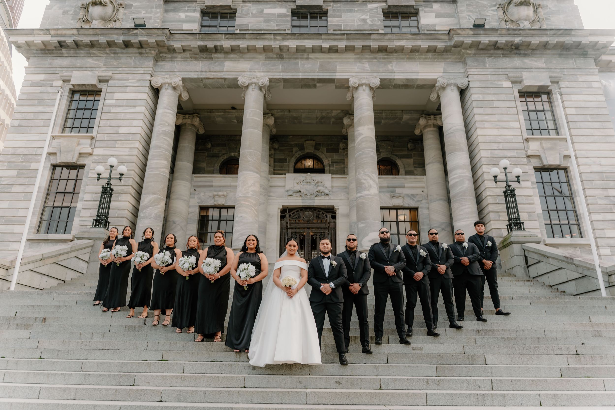 Wedding party poses on grand courthouse steps, bride and groom centered, bridesmaids in black gowns, groomsmen in black suits.