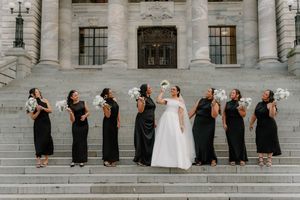 Bride in white gown with bridesmaids in black dresses tossing bouquets on grand stone steps.