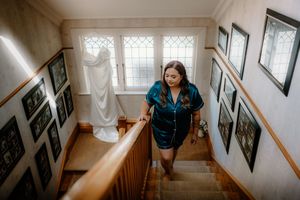 Bride in teal robe descending staircase with wedding dress hanging in background at home.