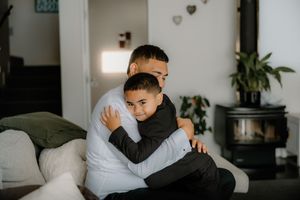 A father and young son share a warm embrace while sitting together in a cozy living room with a fireplace.