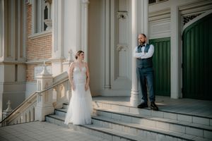 Bride in white gown and groom in navy suit pose on ornate building steps during outdoor wedding photo session.