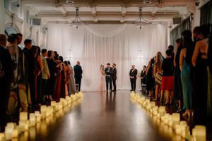 Wedding ceremony in elegant venue with candlelit aisle, couple at altar before white draped backdrop, guests lining both sides.