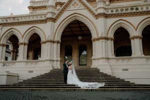 Bride and groom embracing on steps of ornate Gothic-style church with arched columns on their wedding day.