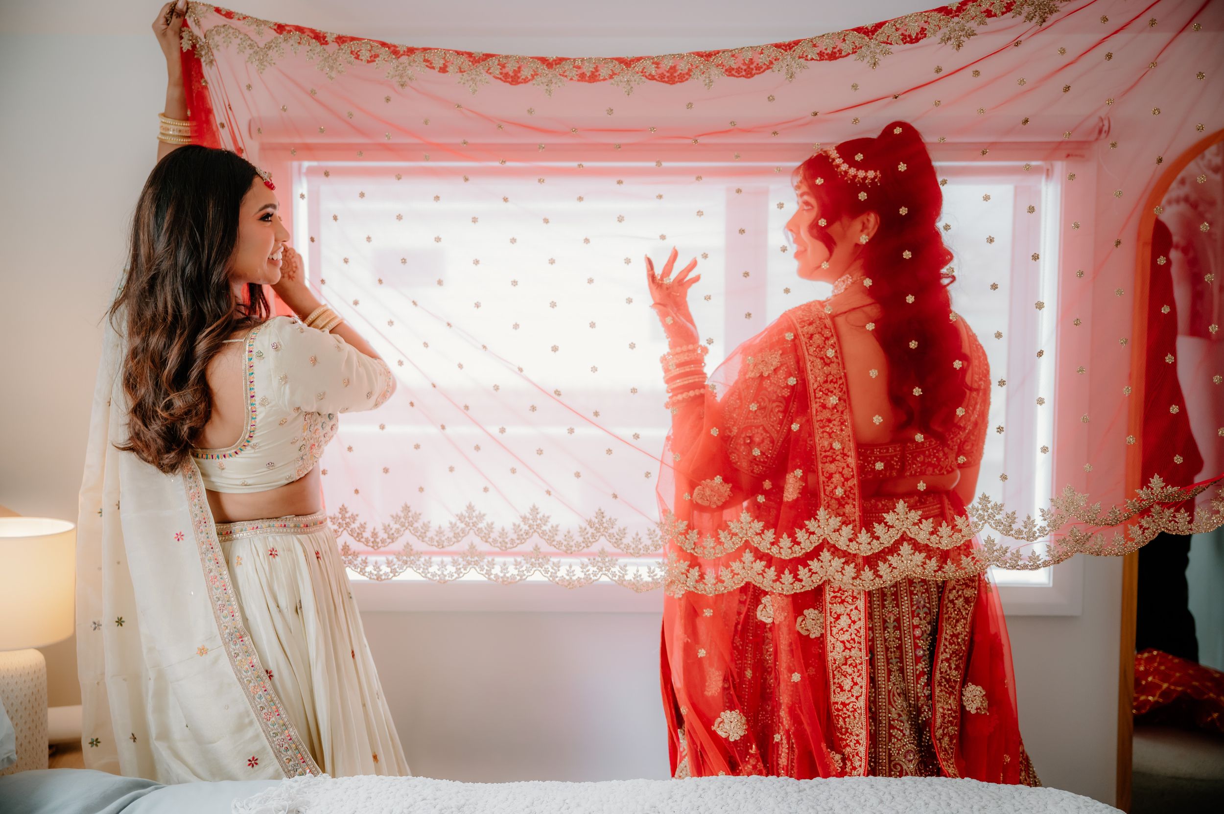 Two women in traditional Indian bridal attire, one in white and one in red lehenga, stand by a window in a bridal suite.