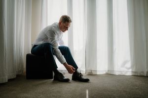 Man in white shirt and jeans sitting on ottoman tying black dress shoes near white curtains.