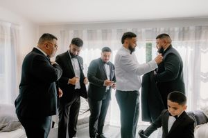 Groomsmen in black tuxedos help each other get dressed before a wedding ceremony in a bright room.