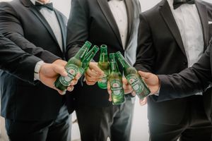 Three groomsmen in black tuxedos toasting with Heineken beer bottles at a wedding celebration.