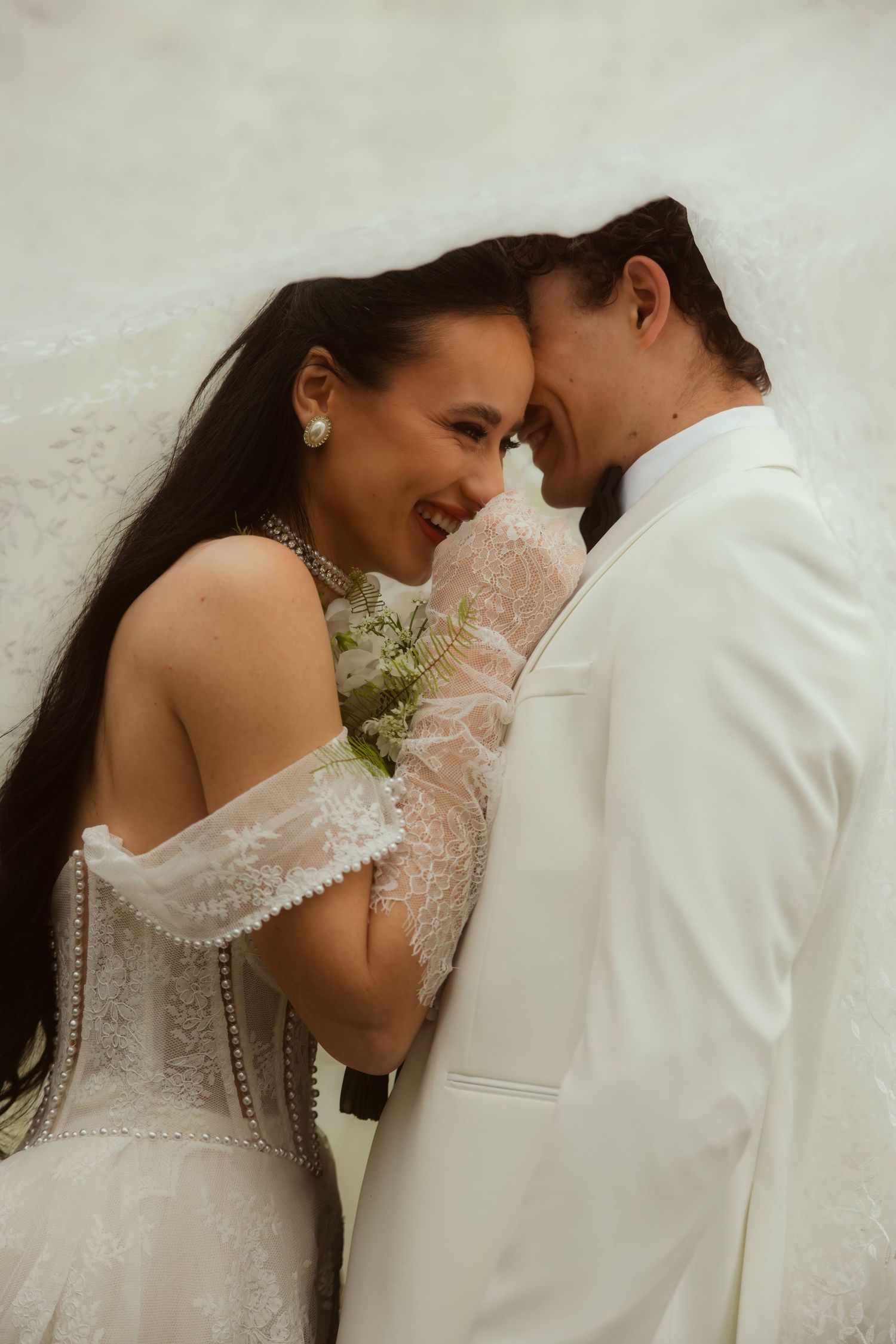 A bride in a lace off-shoulder gown and groom in white suit share an intimate forehead touch while smiling during their Ojai elopement.