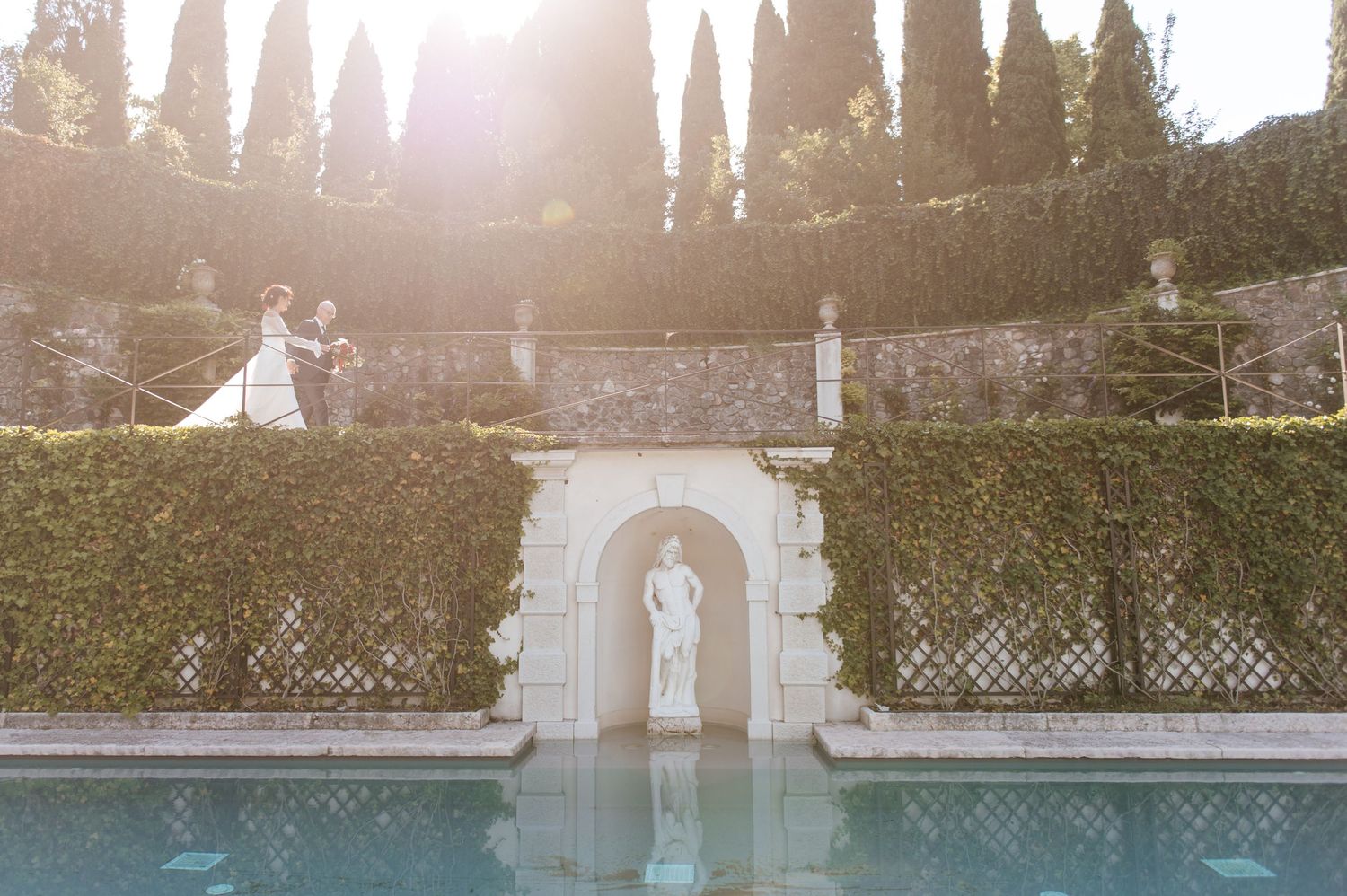 Couple stands atop a lush hedge wall beside a reflecting pool with a classical statue at a sunlit Italian villa.