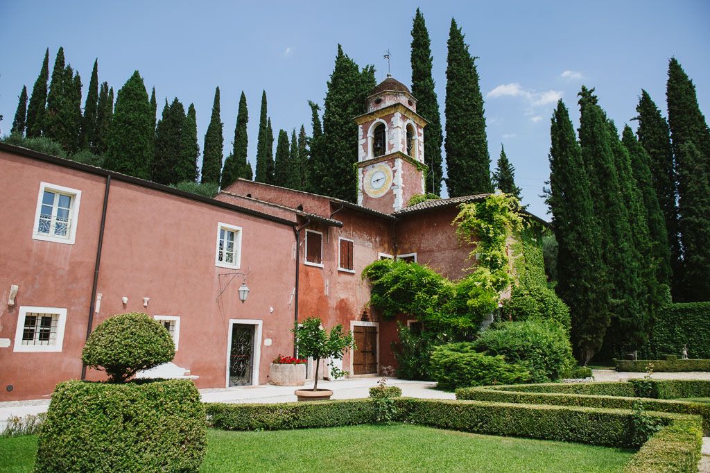 Elegant Italian villa with red facade, clock tower, cypress trees, and manicured hedges under a clear blue sky.