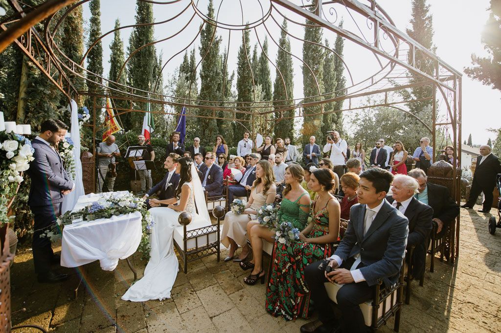 Outdoor wedding ceremony with guests seated under a pergola surrounded by cypress trees in Tuscany.