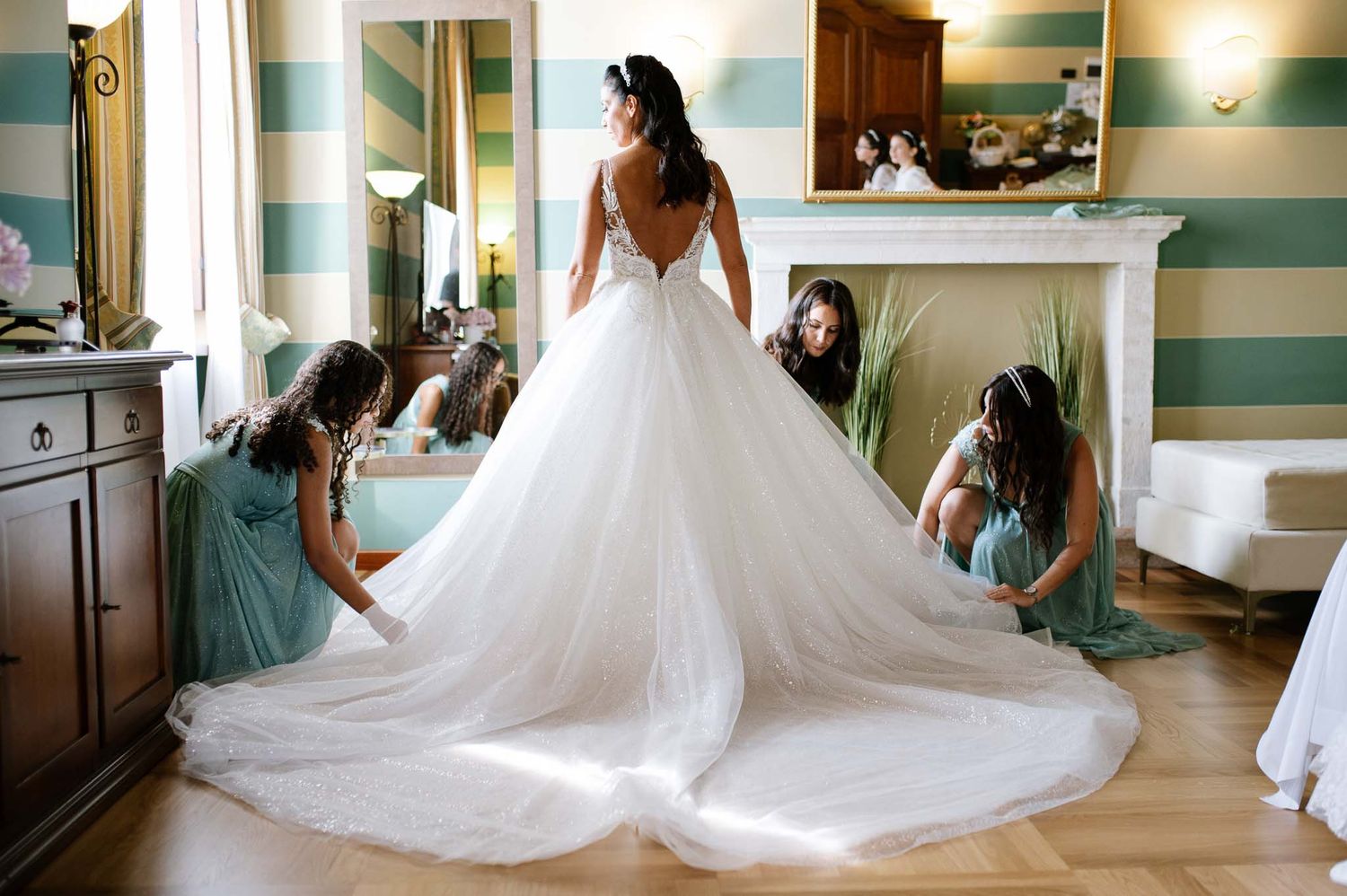 Bride in a stunning backless ball gown wedding dress with long train, assisted by bridesmaids in mint green dresses.