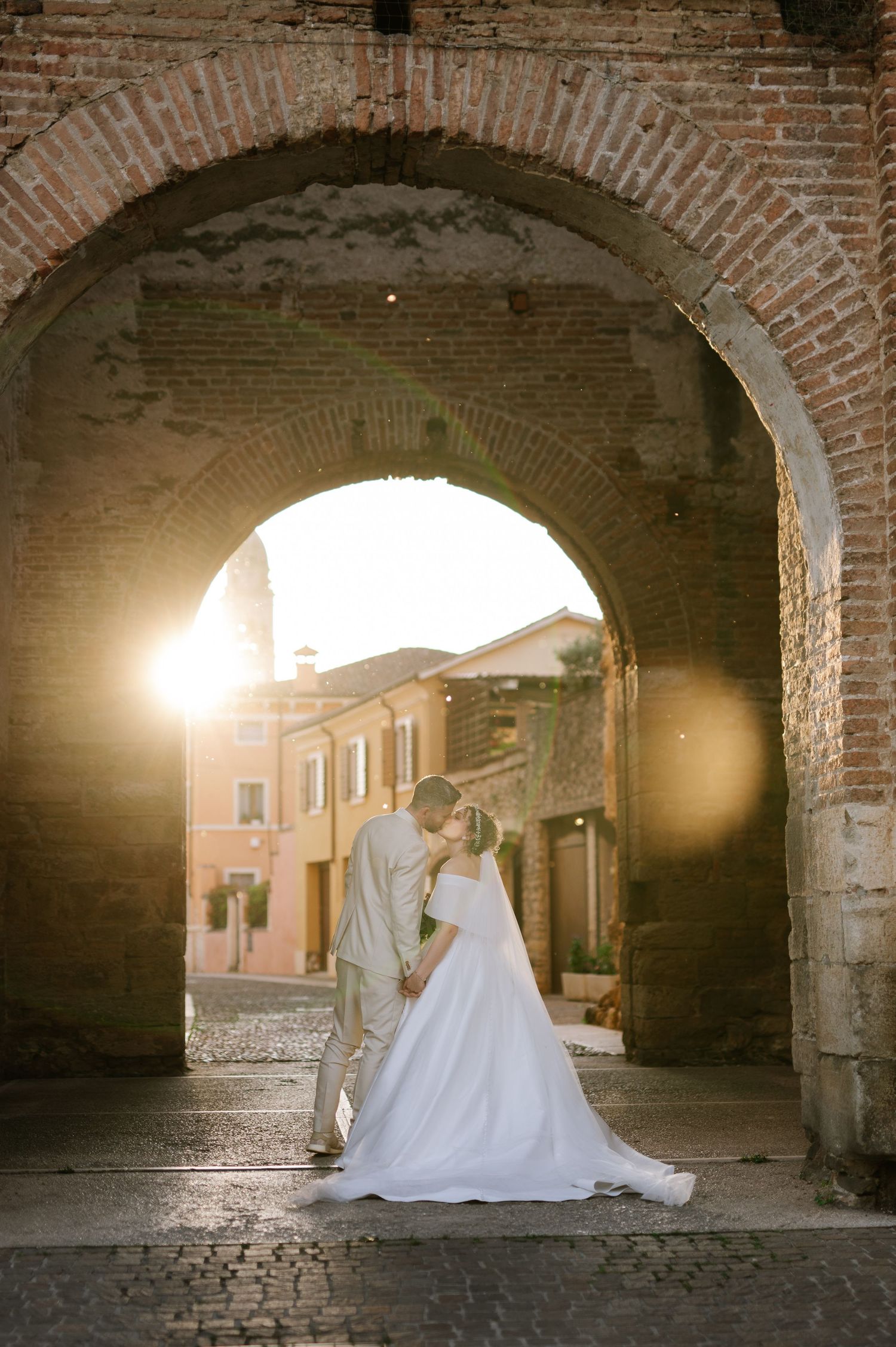 Bride and groom kissing under a rustic brick arch at golden sunset in an Italian cobblestone village.