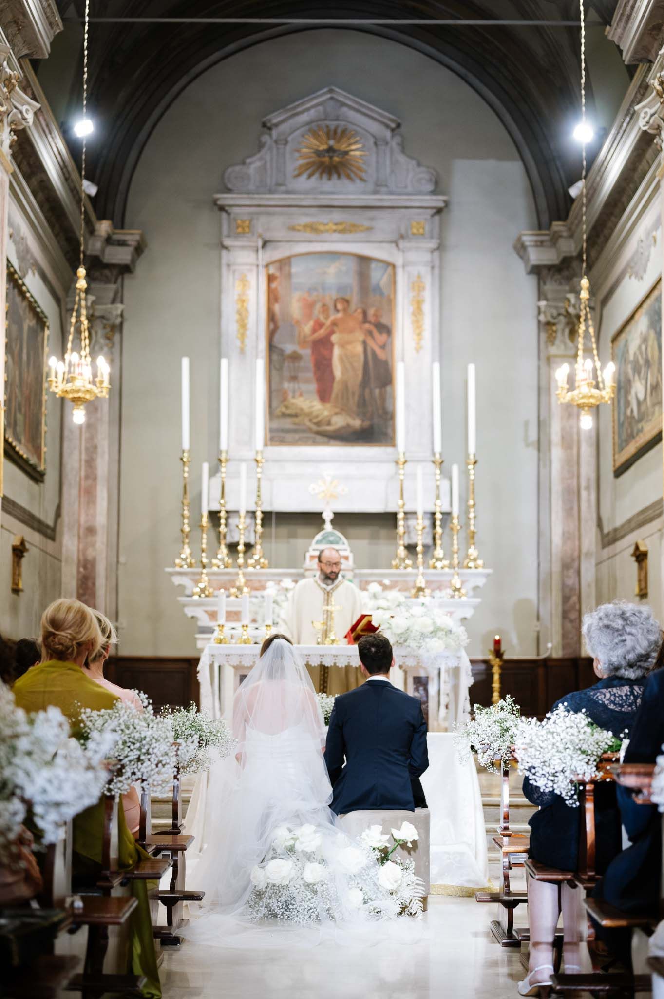 Bride and groom kneeling at ornate church altar during Catholic wedding ceremony with white floral arrangements.