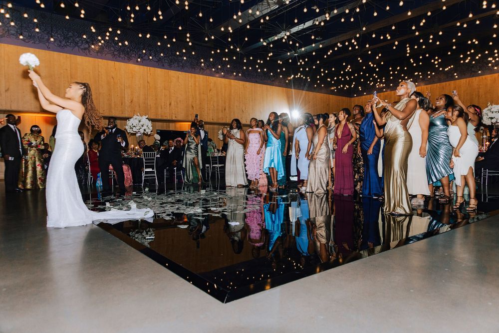 Bride tossing bouquet to guests on reflective black dance floor at elegant wedding reception with twinkling lights.