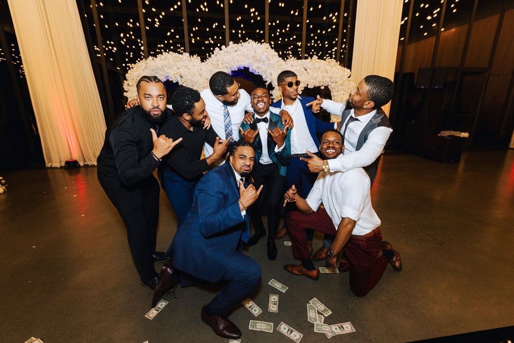 Group of men in suits celebrating at a wedding reception with money on the floor and floral decorations.