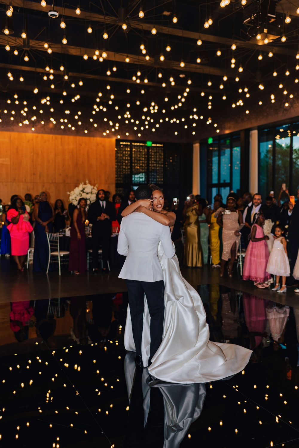 Bride and groom share their first dance embrace on a reflective black floor under warm string lights at elegant wedding reception.