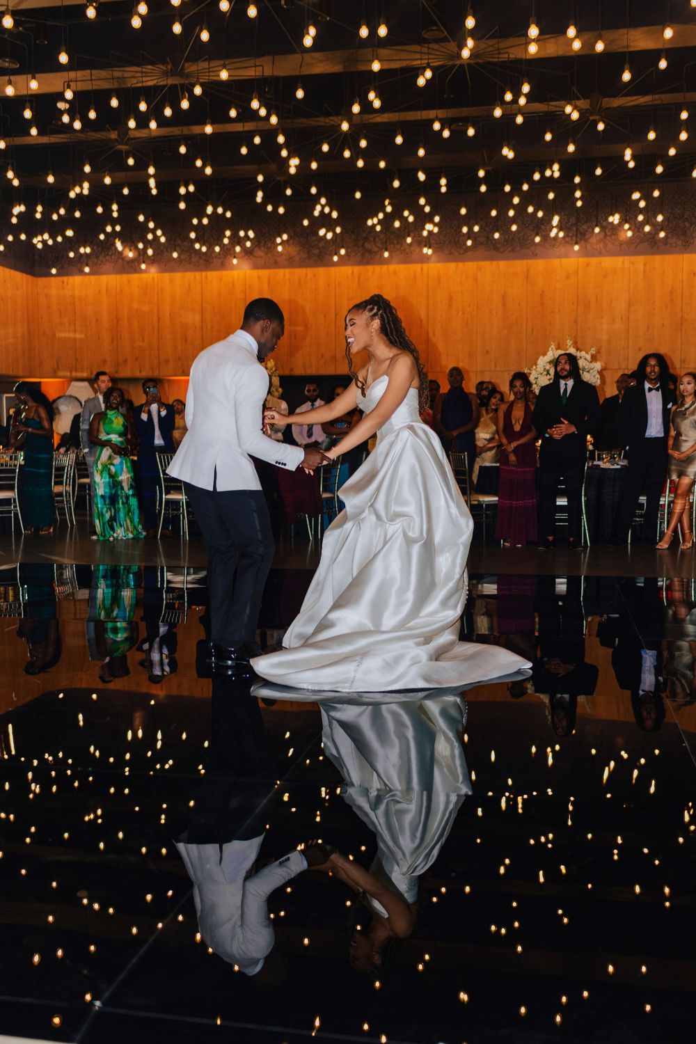 Bride in white satin gown twirls with groom on mirrored dance floor under glowing string lights at wedding reception.