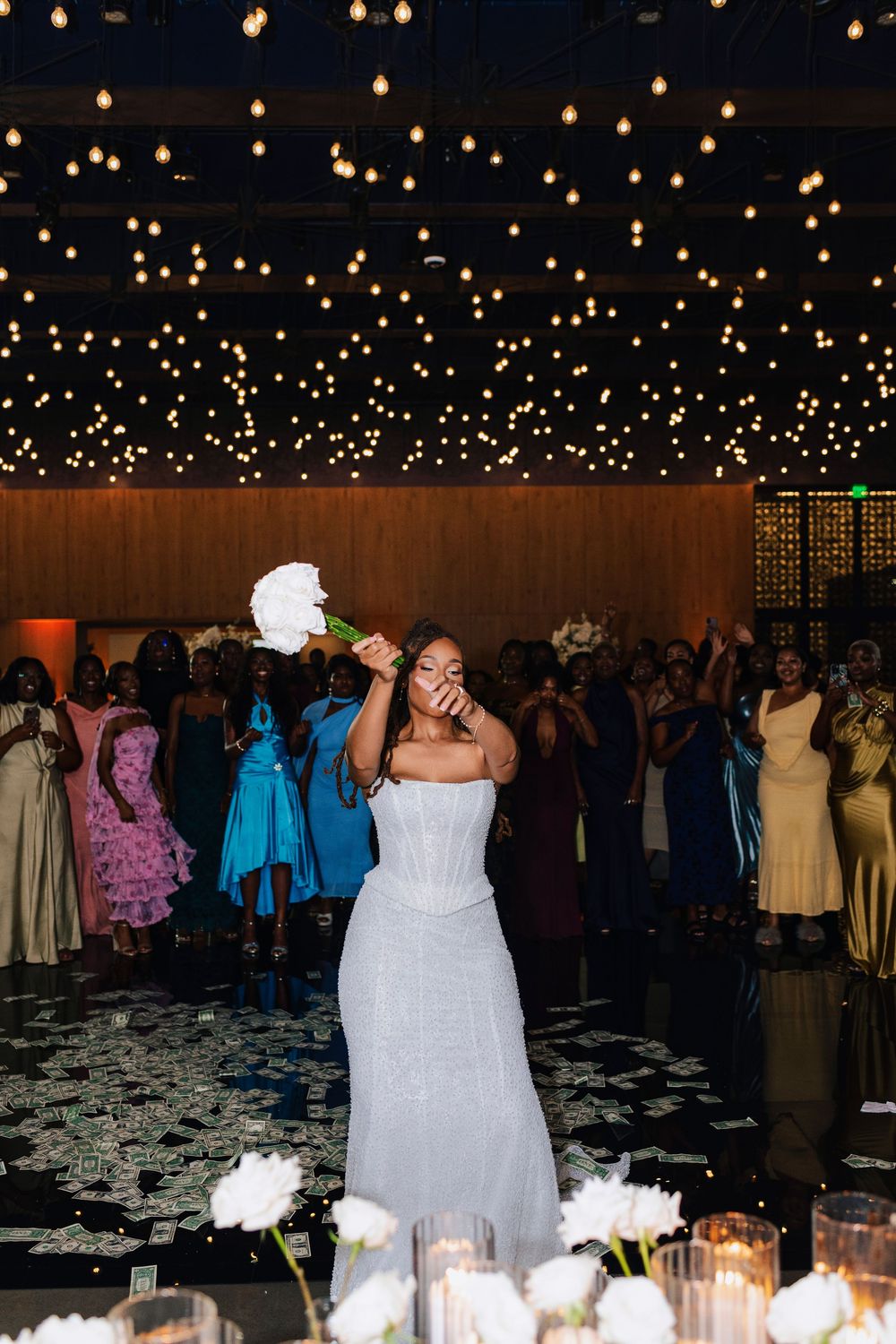 Bride in strapless beaded gown tosses bouquet at wedding reception under twinkling string lights.