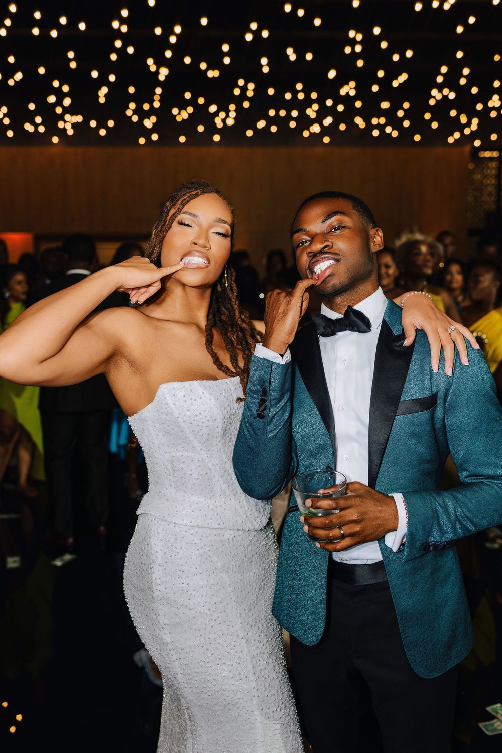 Bride in sparkly white gown poses with groom in teal tuxedo at wedding reception under string lights.