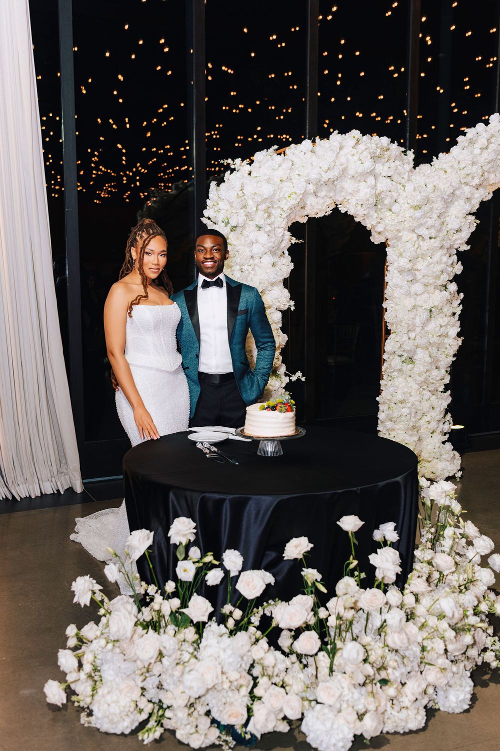 Bride and groom stand beside a white wedding cake surrounded by white floral arrangements and a glowing letter arch.