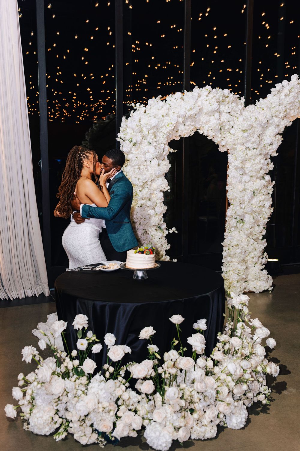 Couple sharing a kiss at wedding cake table beneath white floral arch with roses and fairy lights backdrop.